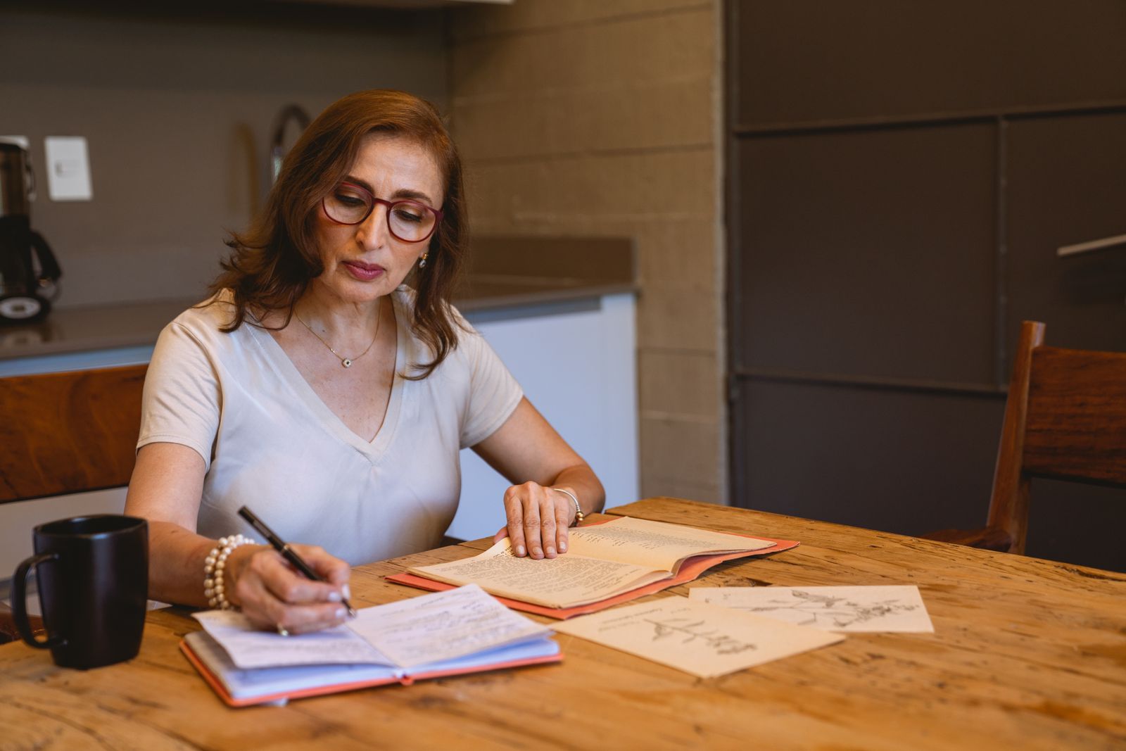 Mulher de óculos escrevendo num caderno sobre a mesa da cozinha, com uma caneca ao lado